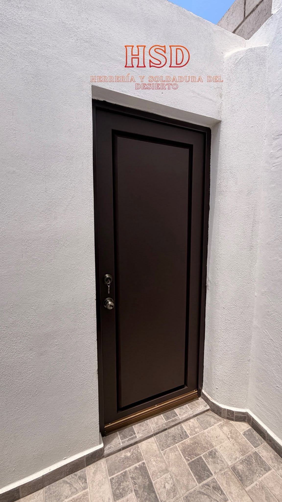 Puerta de acero color café oscuro con tablero liso, instalada en un pasillo de muros blancos