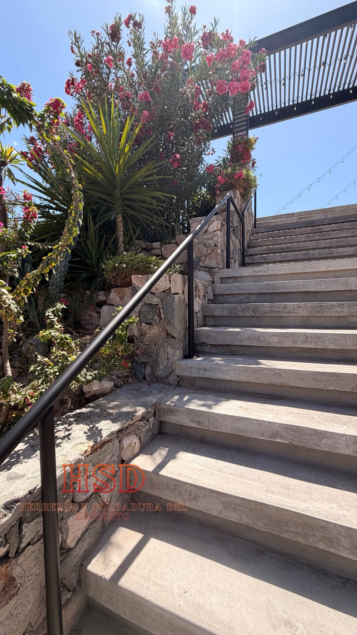 Pasamanos tubular negro a lo largo de una escalera exterior de concreto junto a un muro de piedra con plantas