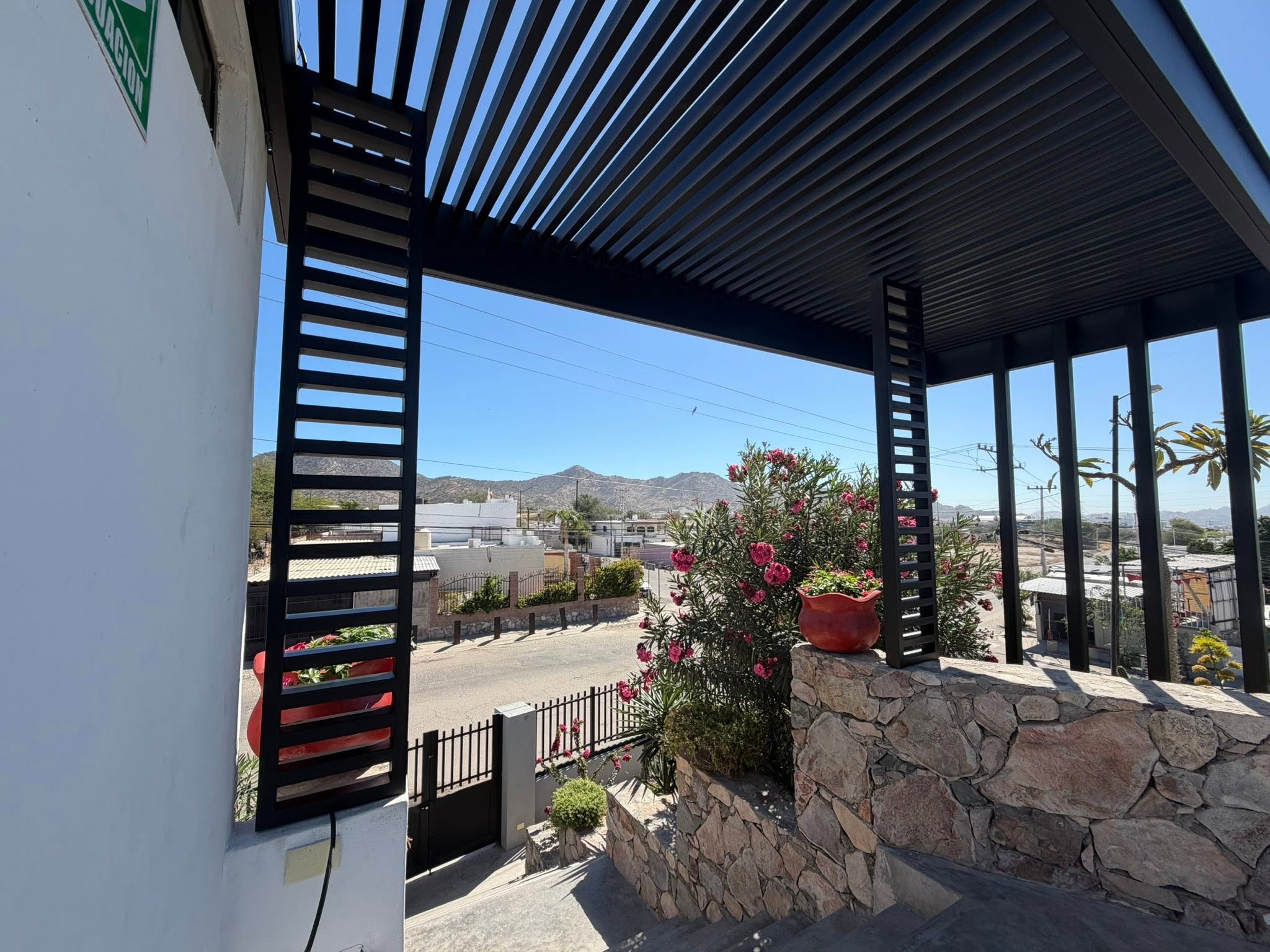 Pérgola de acero negro con techo de lamas y columnas con celosía horizontal sobre una terraza con muro de piedra